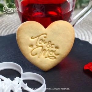 Biscuit en forme de cœur avec l’inscription « Toi & Moi », réalisé à l’aide d’un emporte-pièce Bakers’Tricks, présenté sur une ardoise avec une boisson chaude en arrière-plan.