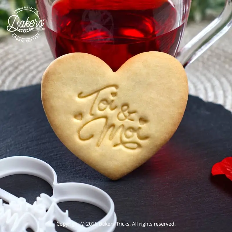 Biscuit en forme de cœur avec l’inscription « Toi & Moi », réalisé à l’aide d’un emporte-pièce Bakers’Tricks, présenté sur une ardoise avec une boisson chaude en arrière-plan.