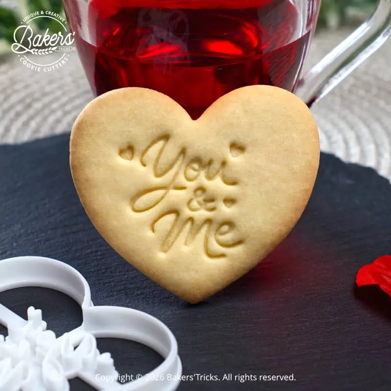 Biscuit en forme de cœur avec l’inscription « You & Me », réalisé à l’aide d’un emporte-pièce Bakers’Tricks, présenté sur une ardoise avec une boisson chaude en arrière-plan.