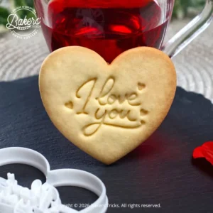 Biscuit sablé en forme de cœur avec l’inscription « I Love You », réalisé à l’aide d’un emporte-pièce Bakers’Tricks, présenté sur une ardoise avec une boisson chaude en arrière-plan.