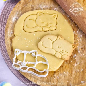 Emporte-pièce Chat endormi Bakers'Tricks découpant une pâte sablée avec le biscuit réalisé juste à côté.